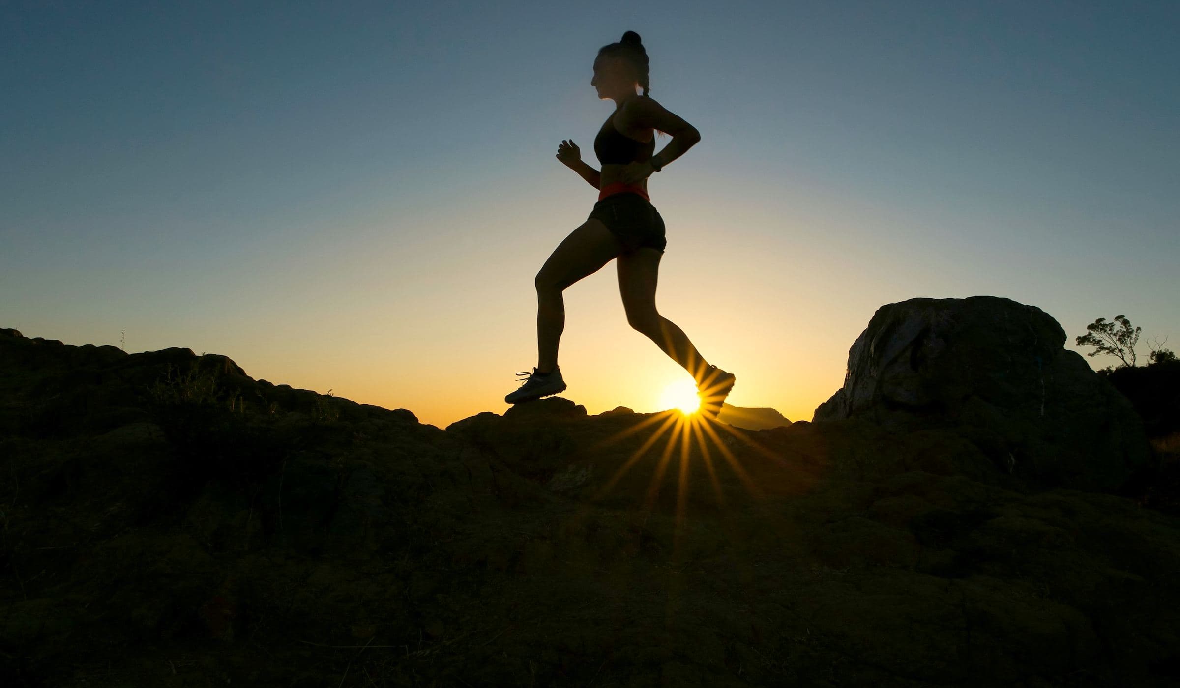 A runner in silhouette striding across a rocky ridge as the sun sets behind her.